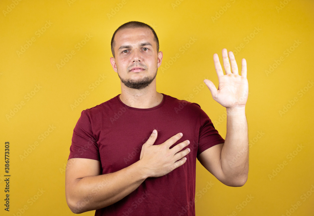 Russian man wearing basic red t-shirt over yellow insolated background smiling swearing with hand on chest and fingers up, making a loyalty promise oath Russian man wearing basic red t-shirt over yellow insolated background smiling swearing with hand on chest and fingers up, making a loyalty promise oath