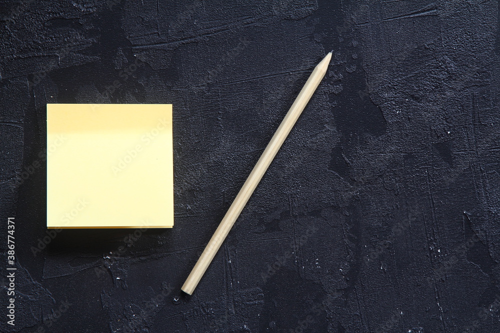 On a black textured rough background, a part of a yellow paper sticker peeks out and a wooden pencil lies on top. On a black textured rough background, a part of a yellow paper sticker peeks out and a wooden pencil lies on top.