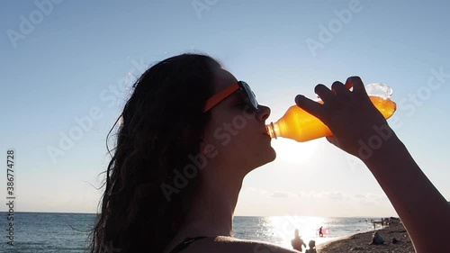 Black silhouette of a girl drinking juice. Young girl drinking water from a plastic bottle against the background of the sea and sky. Teen female having water break at sunset in slowmotion.