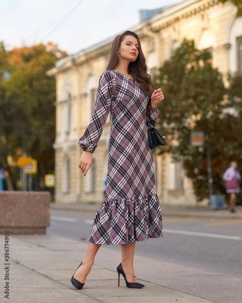 Elegant woman in dress walking city street on fall or autumn day. Pretty girl with long wavy hair holding handbag