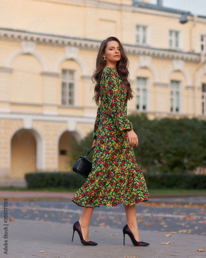Elegant woman in green dress walking city street on fall or autumn day. Pretty girl with long wavy hair holding handbag