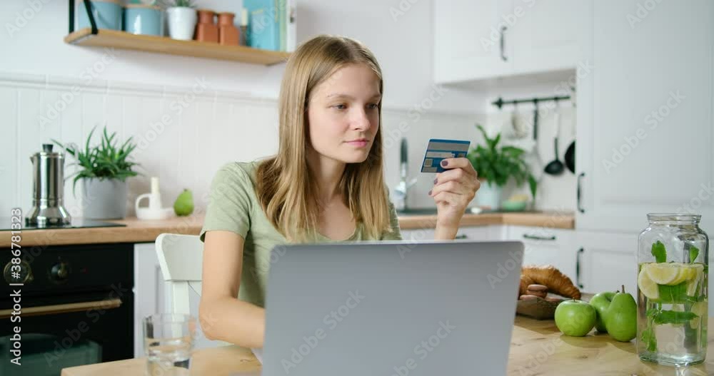 Young Woman Pays Online by Credit Card using Laptop in Kitchen. Customer Buys or makes Purchase at Home in Web Store and Enjoys Shopping via Internet. E-banking and E-commerce Concept. 4K Medium Shot Young Woman Pays Online by Credit Card using Laptop in Kitchen. Customer Buys or makes Purchase at Home in Web Store and Enjoys Shopping via Internet. E-banking and E-commerce Concept. 4K Medium Shot