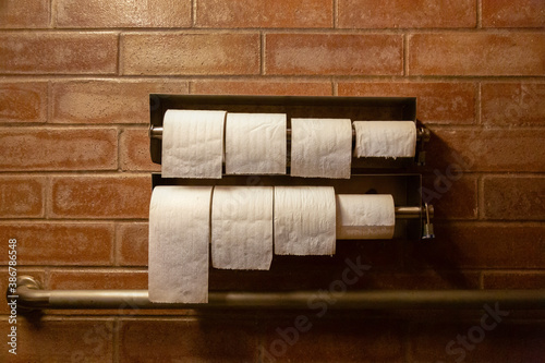 An eye level view of multiple rolls of toilet paper in a metal roll holder mounted on a brick wall with a metal handicap bar below 