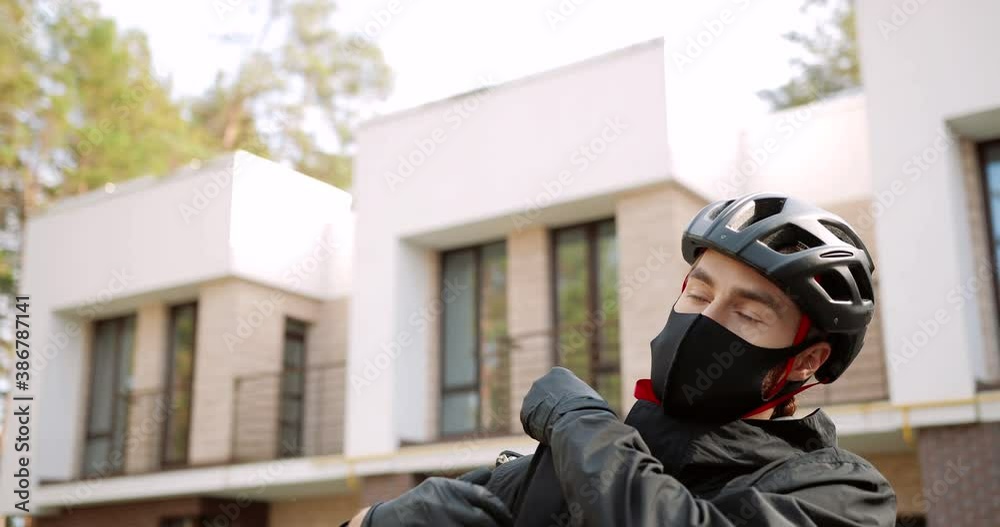 Close up of Caucasian young male courier taking thermal backpack from ground and walking on street. Handsome delivery man in mask and helmet outdoors in town. Working in quarantine. Supply concept Close up of Caucasian young male courier taking thermal backpack from ground and walking on street. Handsome delivery man in mask and helmet outdoors in town. Working in quarantine. Supply concept