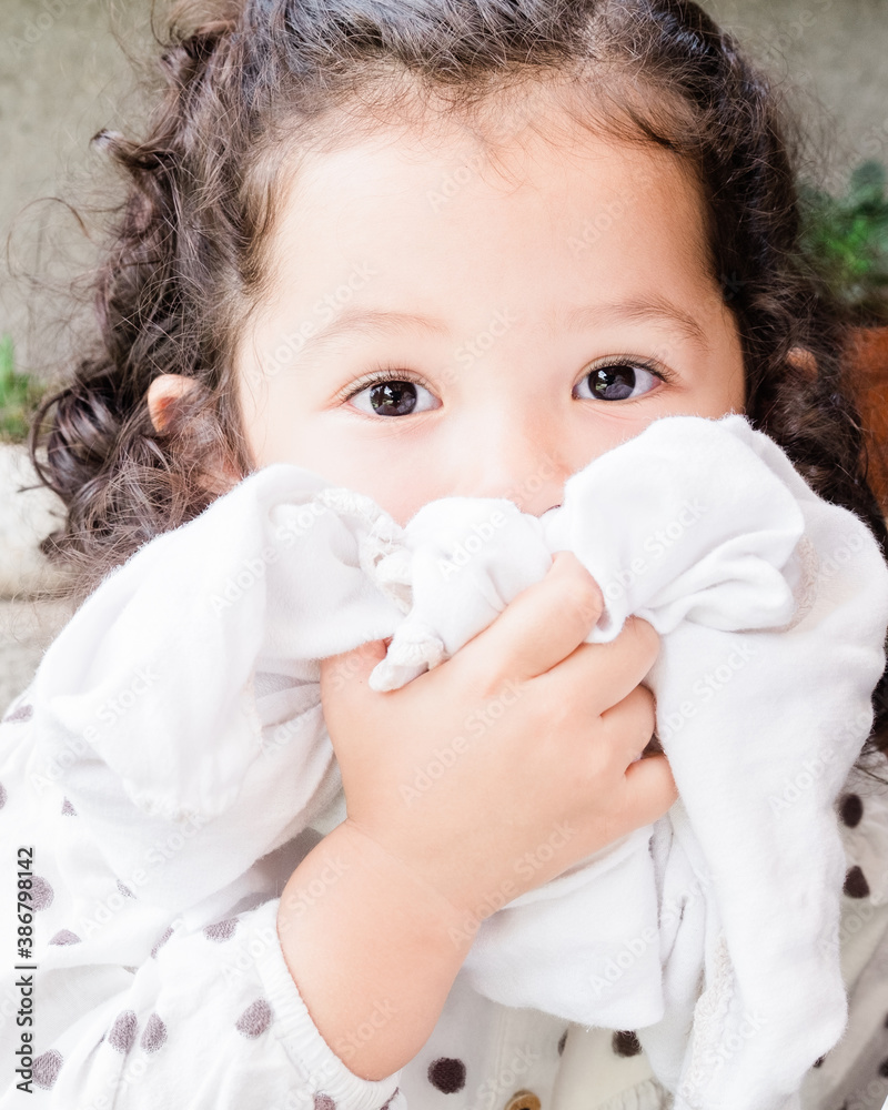 Hispanic toddler with big eyes covering her face with a blanket