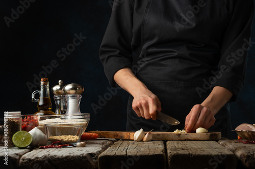 The professional chef in black uniform cuts with knife garlic on chopped wooden board at rustic table with ingredients for cooking background. Backstage of preparing meal with vegetables.