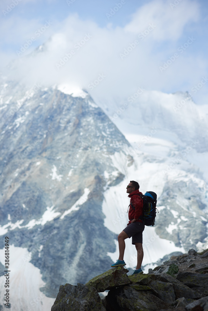 Vertical snapshot silhouette of tourist with backpack standing on big stone, looking at beautiful mountains with snow. Trekking, mountain hiking. Wild nature with amazing views. Sport tourism in Alps.