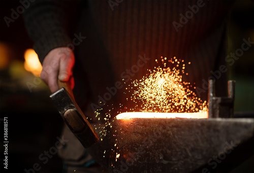 The blacksmith hits the red-hot workpiece in the forge with a hammer and glowing sparks fly in all directions