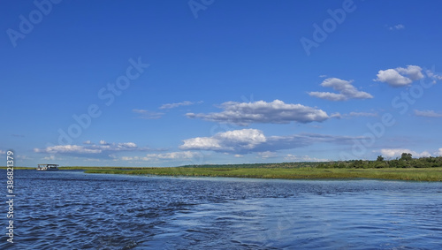 Peaceful summer landscape. The blue river flows between green banks. There are clouds in the azure sky. The ship is visible in the distance. Botswana. Chobe.