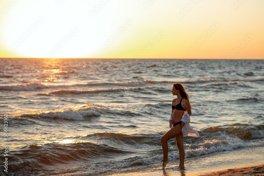 A young beautiful woman in a black bikini and a white shirt on a tanned body is walking along the beach at sunset. Soft selective focus, art nose.