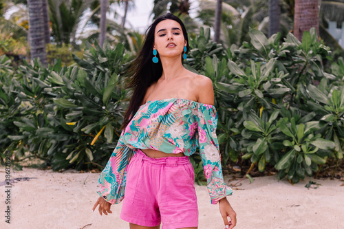 Fashion portrait of stylish woman in colorful print long sleeve top and pink shorts on beach, tropical background.  