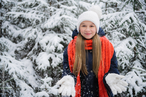 A young teenage girl in a down jacket smiles beautifully for a picture in a winter snow covered forest.