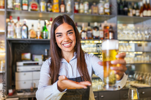 Beautiful smiling female Bartender serving a draft beer at the bar counter , shelves full of bottles with alcohol on the background