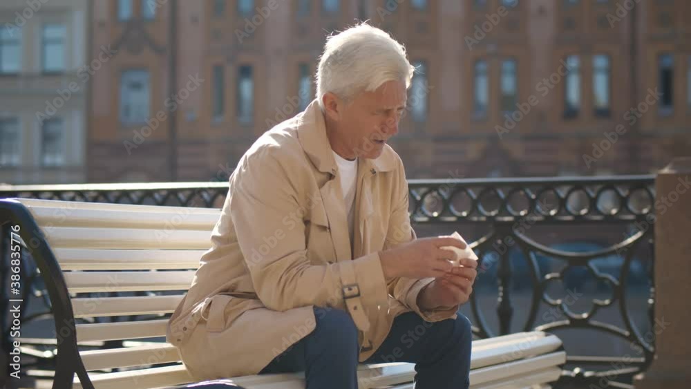 Side view of senior man sitting on bench and sneezing in paper tissue having flu