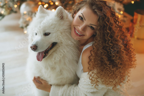 Beautiful woman sits on a white blanket with a dog hugs, against the background of a Christmas tree fireplace with candles in a decorated room. Happy New Year and Merry Christmas. High quality photo. © nuzza11