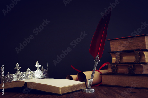 low key image of beautiful queen/king crown, old books and feather quill ink pen over wooden table. fantasy medieval period