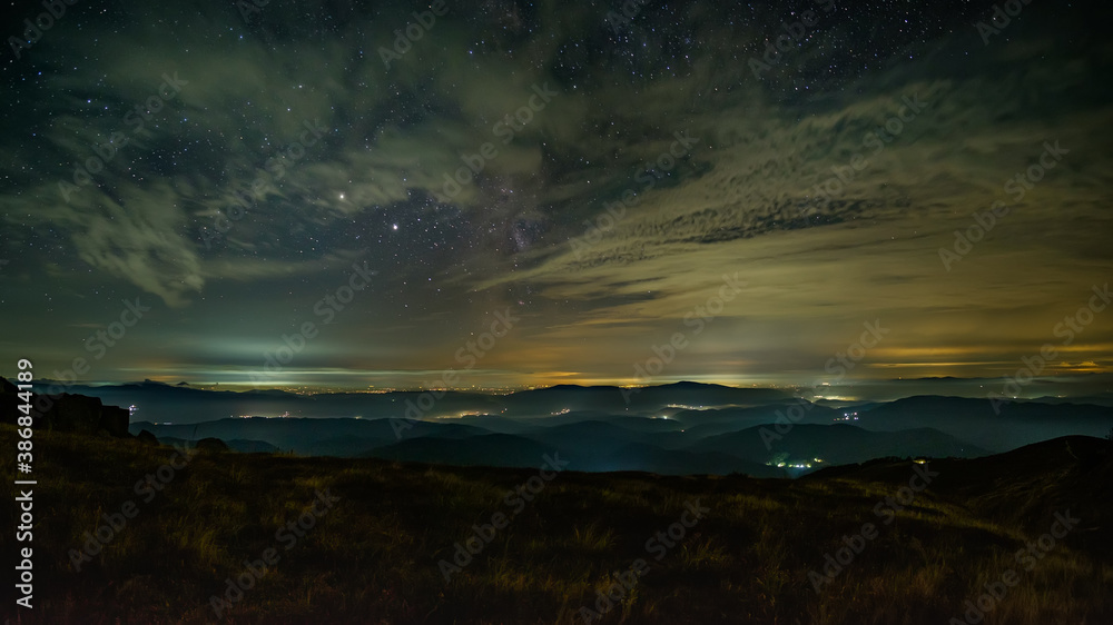 Night panorama of the mountainous area. Small villages glow in the night. Low clouds reflect light. The stars sparkle between the clouds