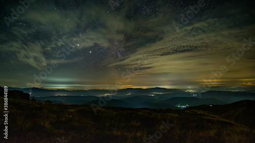 Night panorama of the mountainous area. Small villages glow in the night. Low clouds reflect light. The stars sparkle between the clouds