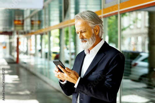 Portrait of senior man in business suit using phone in front of glass building in the city