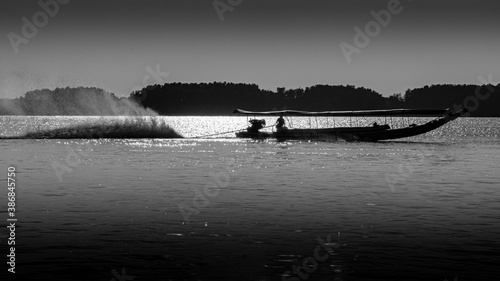 Black and white silhouette of Thai long tail speed boat is running in the sea. The high speed of fan behind the boat creates big and long water splash on the water surface.
