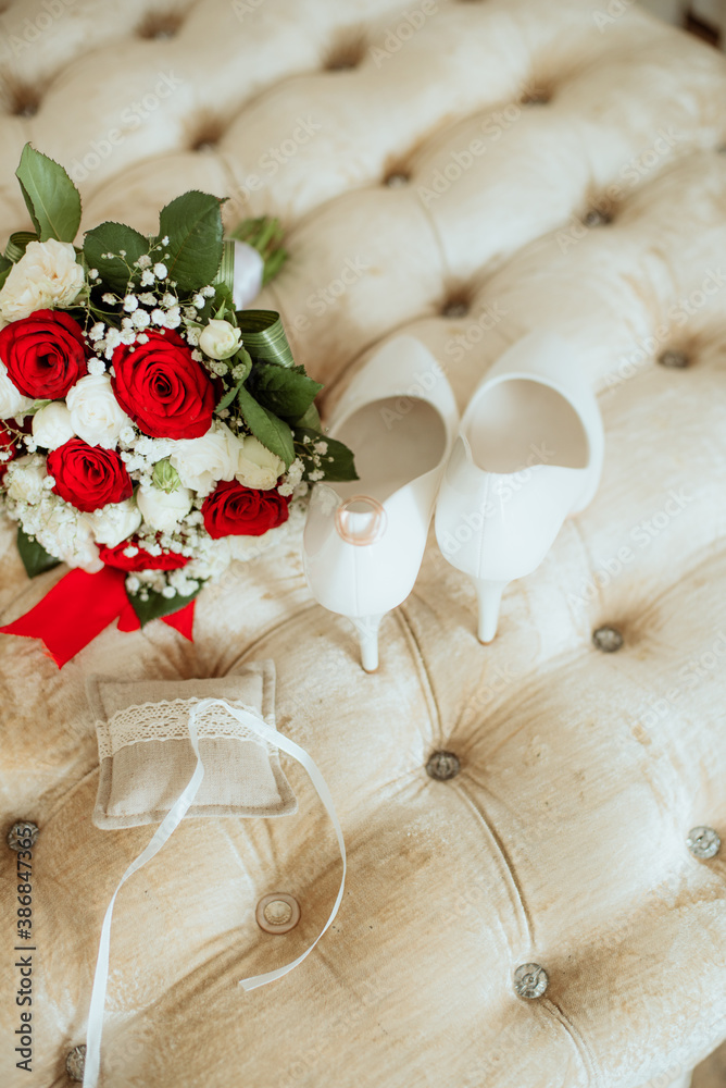 Wedding bouquet of white and red roses, white high heels and gold wedding rings on a boudoir ottoman. Wedding details