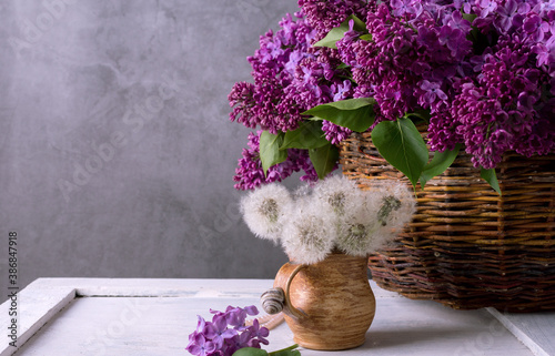 Fotografie Beautiful dandelion bouquet in a brown ceramic pot against purple lilac flowers in dark moody style