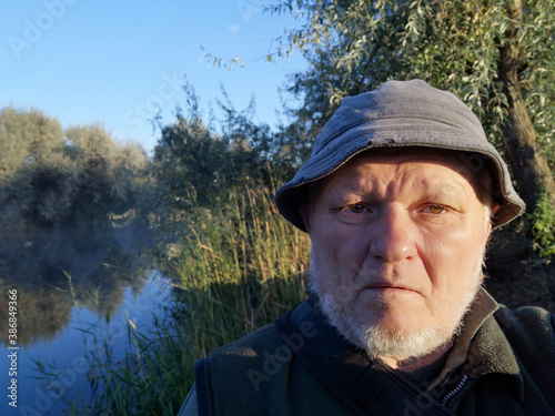 An elderly man with gray beard in hat on background of forest and river