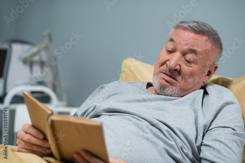 A male patient in his mid sixties reading a book in a hospital ward.