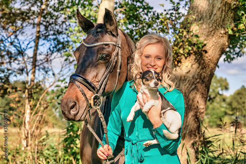 Beautiful portrait of a blond smiling girl with her horse and dog in the forest. Wearing a green dress. Selective focus © Dasya - Dasya