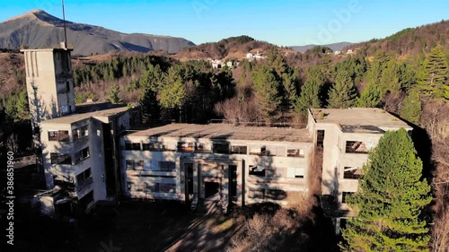 Aerial footage of an abandoned building in the trees, with mountains in the background and a blue sky - Colonia di Rovegno, Italy photo