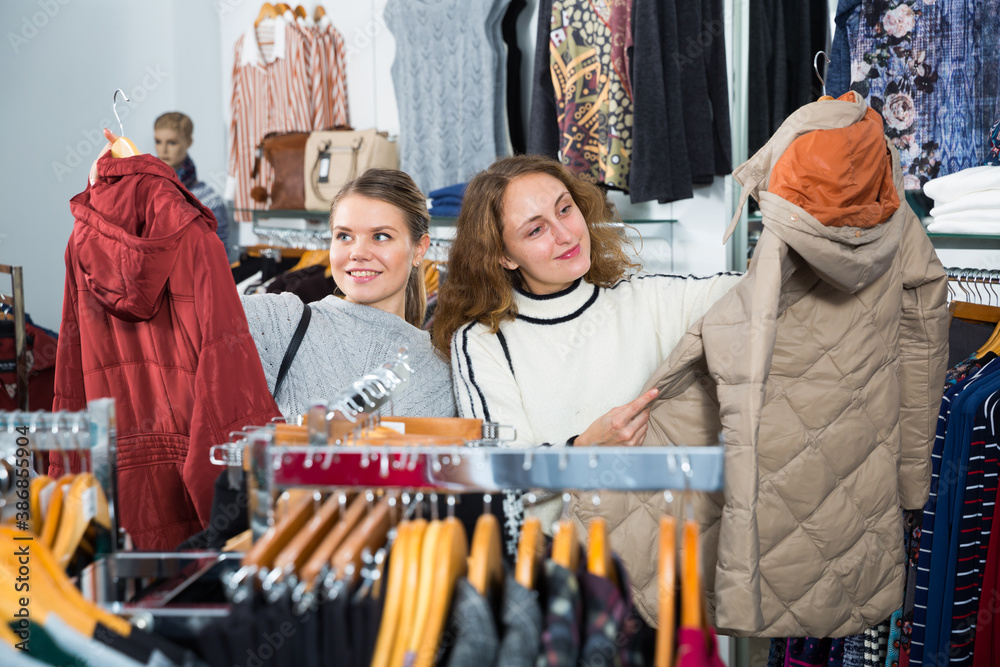 Portrait of two cheerful female friends choosing new coat in showroom