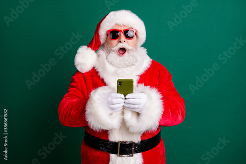 Photo of pensioner stylish grandpa grey hair beard hold telephone shocked open mouth crazy novelty wear red santa x-mas costume coat gloves sunglass headwear isolated green color background