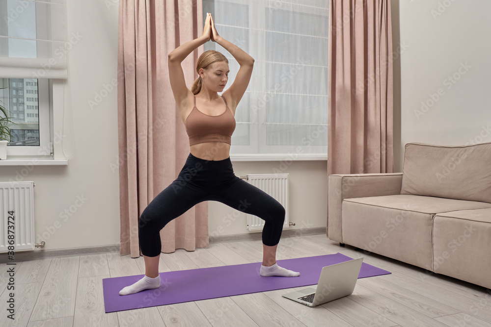 A young woman learns yoga online on the Internet, looks at the screen in a laptop, poses in yoga, on the floor there is a laptop with an open screen.