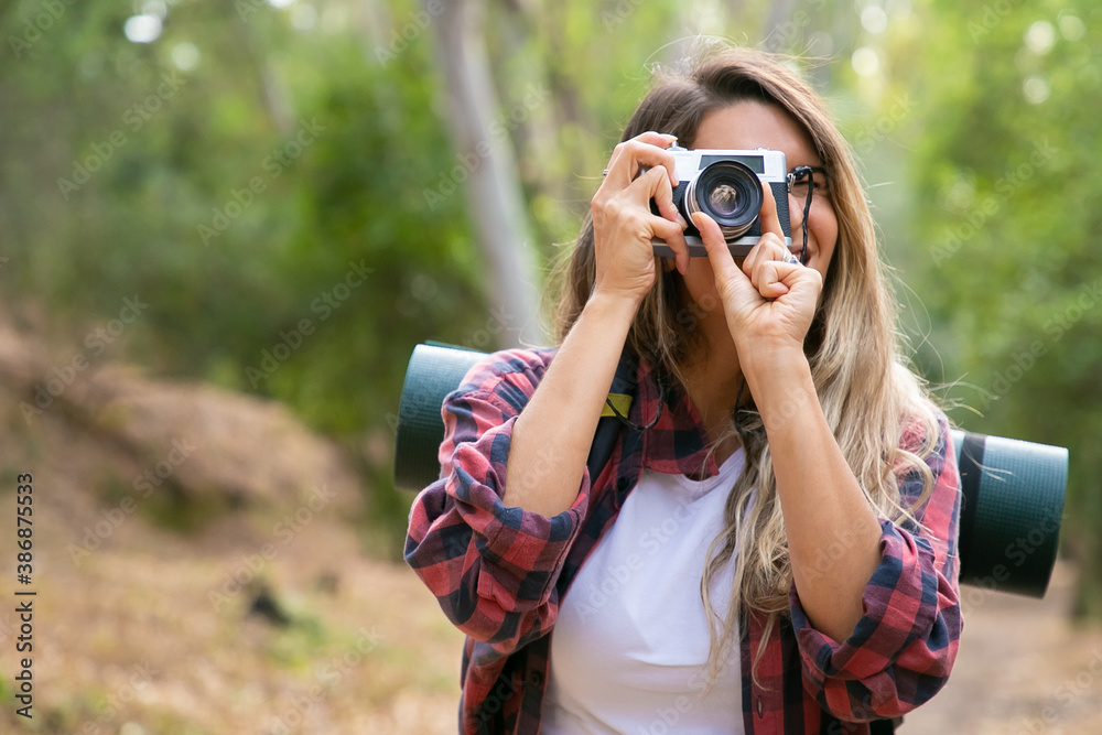 Happy blonde woman taking photo of nature with camera and smiling. Caucasian long-haired traveler walking or hiking in forest. Blurred background. Tourism, adventure and summer vacation concept
