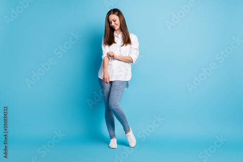 Full size photo of pretty lady long hairstyle confident person fixing sleeve white shirt jeans sneakers isolated blue color background