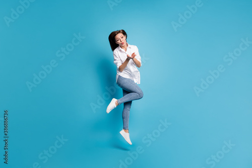 Full length photo of pretty lady jump high up sending air kisses wear white shirt jeans shoes isolated blue color background