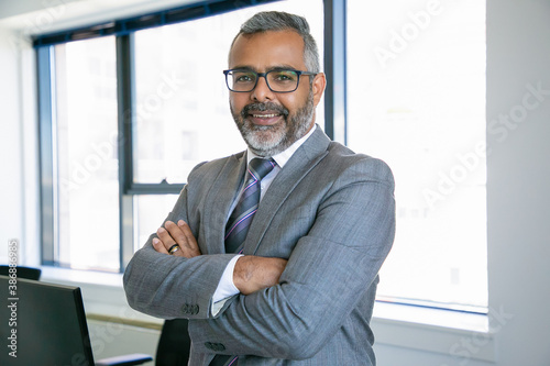 Experienced businessman standing in office room and looking at camera. Indian content office employee in eyeglasses smiling and posing with folded hands. Business, management and corporation concept