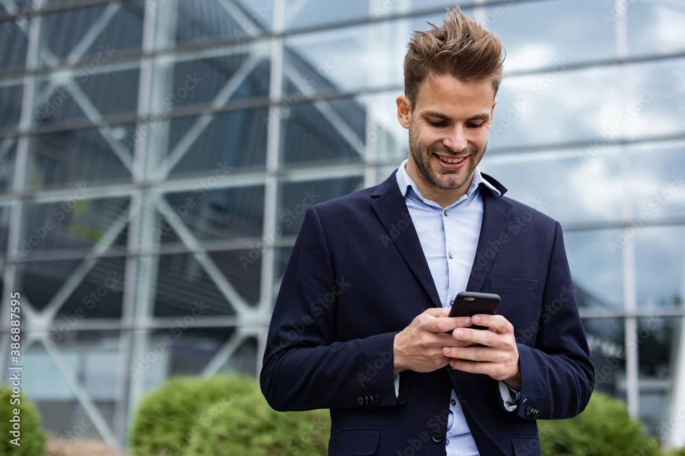 Handsome office worker holding smartphone and smiling. Happy young man using mobile phone apps, texting message, browsing internet, looking at smartphone. Coffee break near the business center. Handsome office worker holding smartphone and smiling. Happy young man using mobile phone apps, texting message, browsing internet, looking at smartphone. Coffee break near the business center.