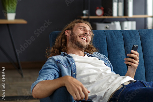 Curly-haired guy in casual style, watching great news on his mobile phone. Student receiving sms message reading good news. Surprised Man celebrating Victory on phone in Apartment.