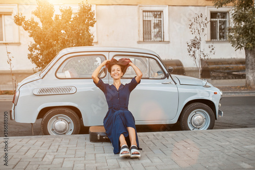 Smiling female posing near stylish old fashioned car. Woman sitting on suitcase. Street style and city walk concept