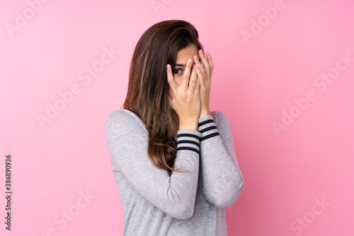 Teenager girl over isolated pink background covering eyes and looking through fingers © luismolinero