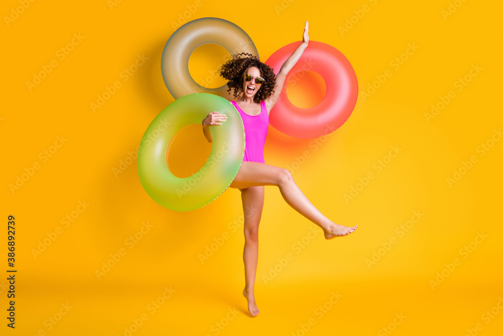 Photo portrait of excited girl standing on one leg wearing pink swim wear holding green red blue inflatable ring isolated on vivid yellow colored background - obrazy, fototapety, plakaty 