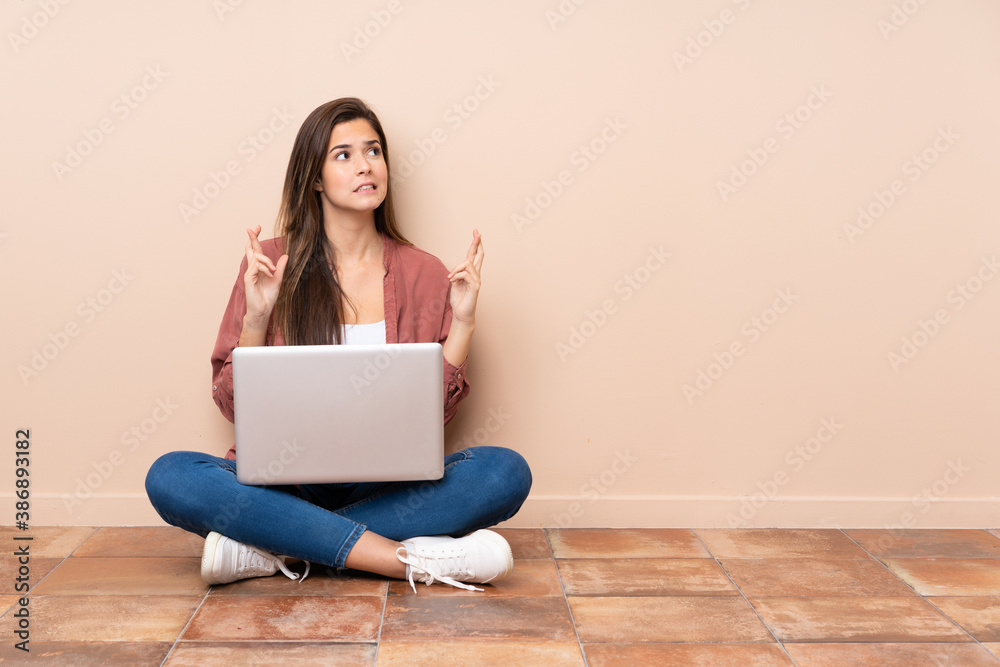 Teenager student girl sitting on the floor with a laptop with fingers crossing and wishing the best Teenager student girl sitting on the floor with a laptop with fingers crossing and wishing the best