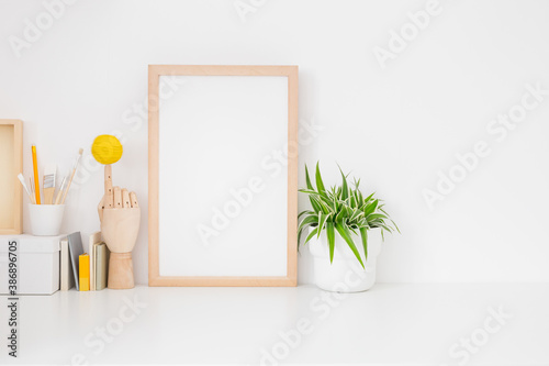 Wooden frame mockup. Creative desk, home workspace, with wooden supplies, plant, hand model, and white wall background.