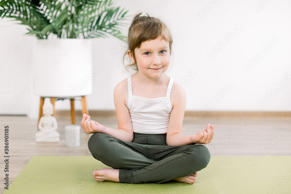 Little Caucasian kid girl wearing sportswear, meditating in a lotus pose with mudra gesture, sitting on green mat, at cozy yoga studio or home. Children doing yoga Little Caucasian kid girl wearing sportswear, meditating in a lotus pose with mudra gesture, sitting on green mat, at cozy yoga studio or home. Children doing yoga