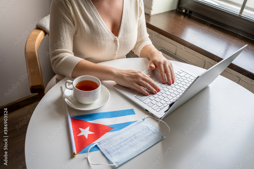 Woman tourist having breakfast with cup of coffee working on laptop.  Flag of Cuba, medical protective face mask on table of cafe. Protection from bacteria and viruse in a public place. Concept.