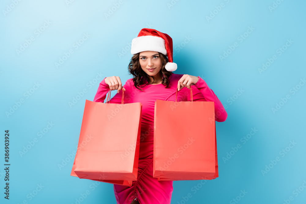 young brunette woman in santa hat holding shopping bags and looking at camera on blue young brunette woman in santa hat holding shopping bags and looking at camera on blue