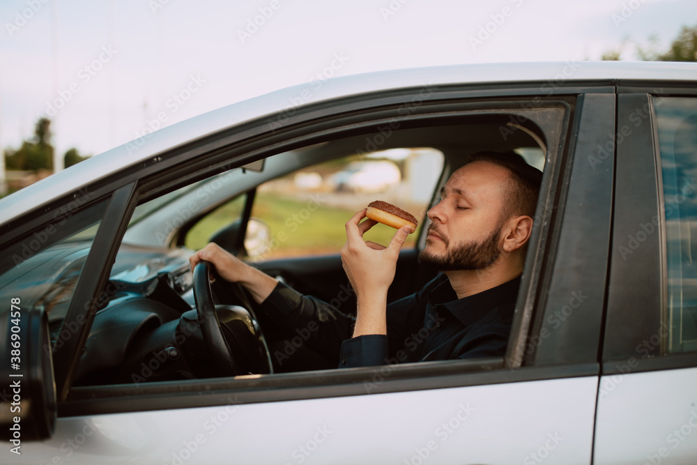 Side view of a caucasian businessman holding a sweet doughnut with chocolate in his hand and driving a car on the way to work. A young man's meal stuck in traffic