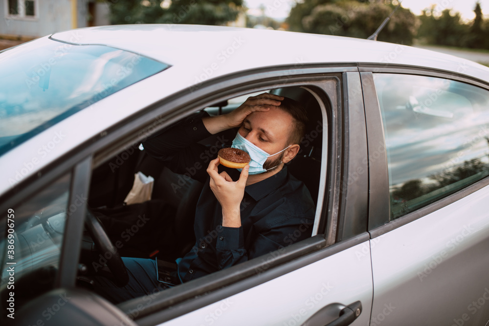 A portrait of a young man with a mask on his face who is on a diet while sitting in the car and looking at the donut he is holding in his hand. Protection against COVID - 19 coronavirus