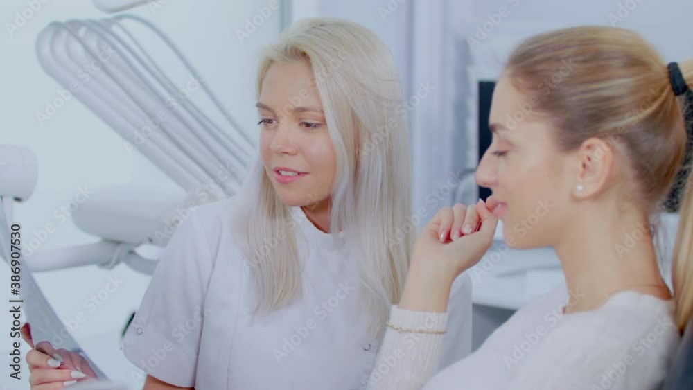 Professional Blonde Female Dentist Shows Patient The Situation Of Teeth X-Ray Image Discussion Of The Treatment Plan And Healthy Smile. Concept Of Clean And Healthy Teeth Slow Motion
